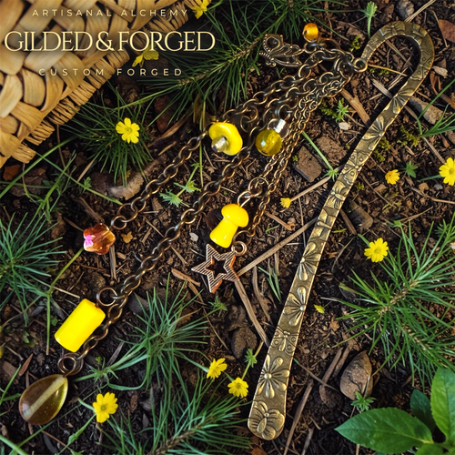 Antique brass bookmark with gold star charm, crystal flower charm, mushroom charm, and yellow glass beads resting on forest floor with small yellow wildflowers. Gilded and Forged in upper left corner.