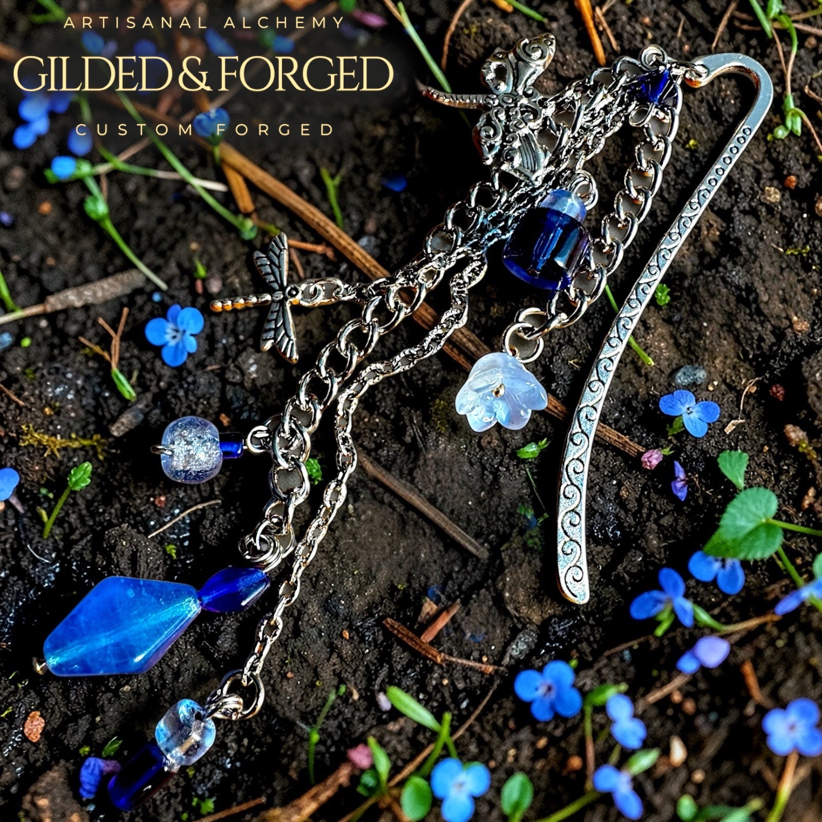 Antique silver dragonfly charm bookmark with cascading chain and deep blue glass crystal beads resting on dark soil with small blue flowers, handcrafted by Gilded & Forged.