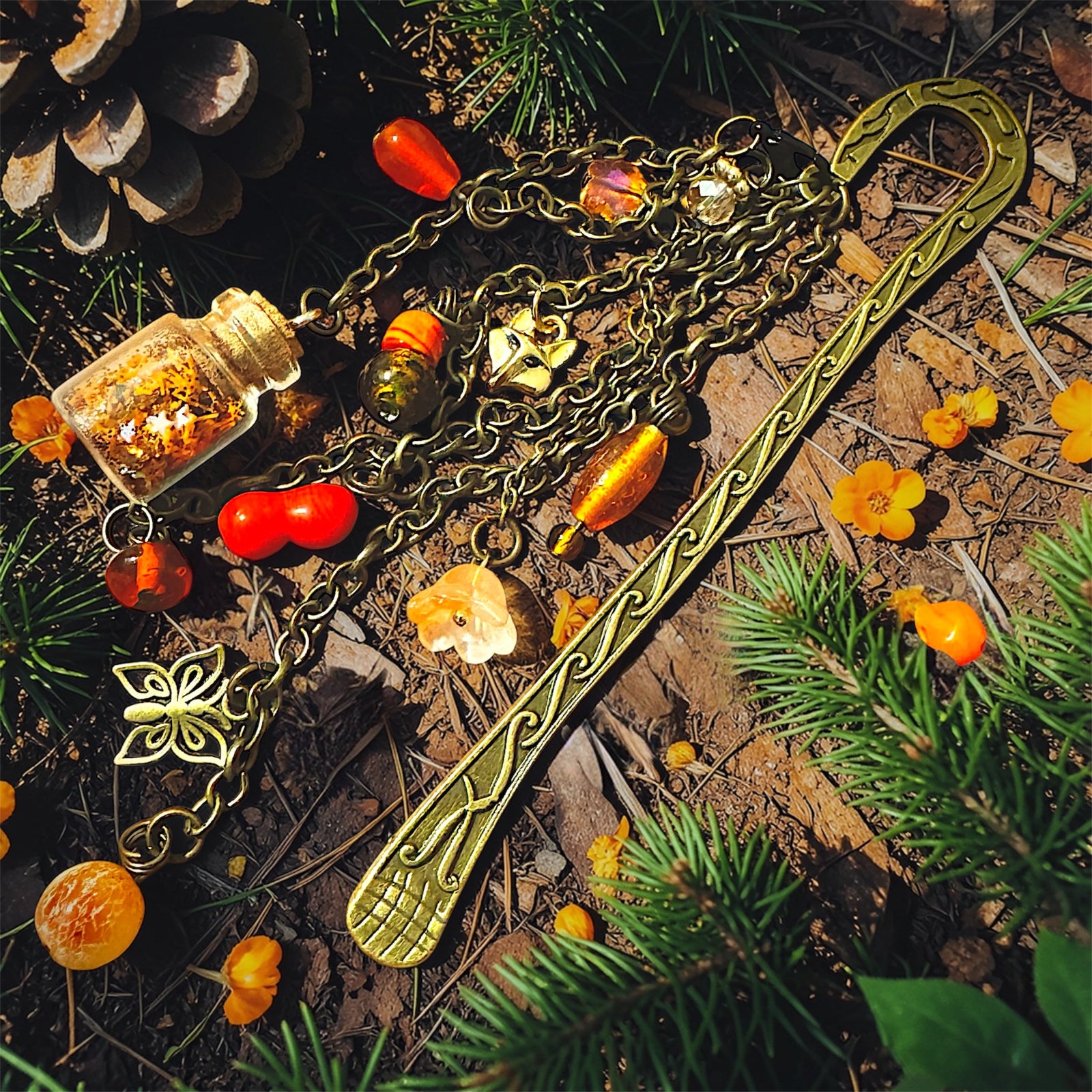 Antique brass fox charm bookmark with butterfly charms, flower charm, amber glass beads, and tiny corked bottle charm resting on forest floor with pine needles.
