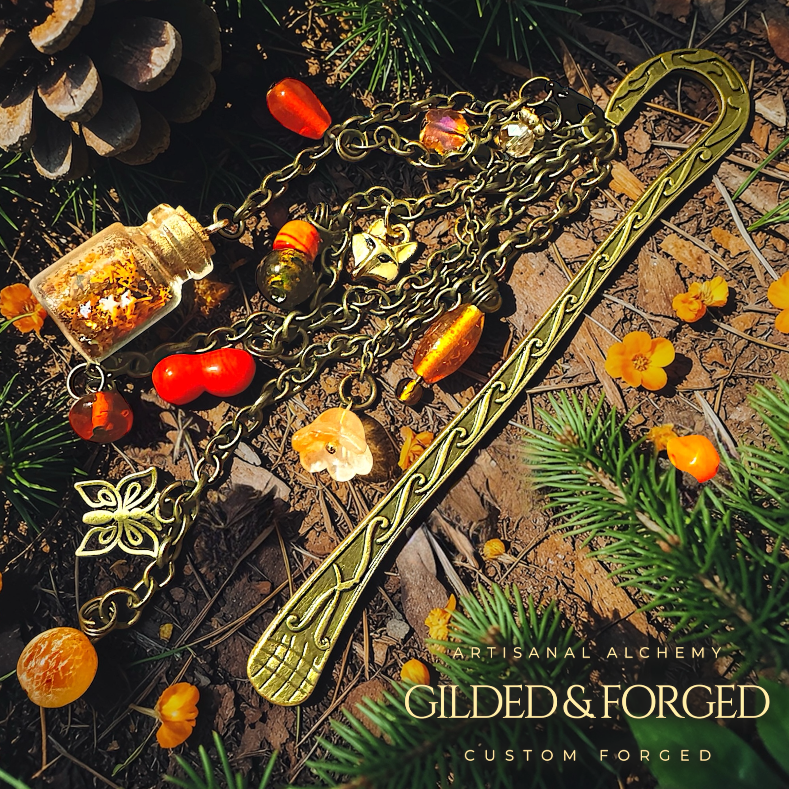 Antique brass fox charm bookmark with butterfly charms, flower charm, amber glass beads, and tiny corked bottle charm resting on forest floor with pine needles. The words Gilded & Forged in the lower right corner.