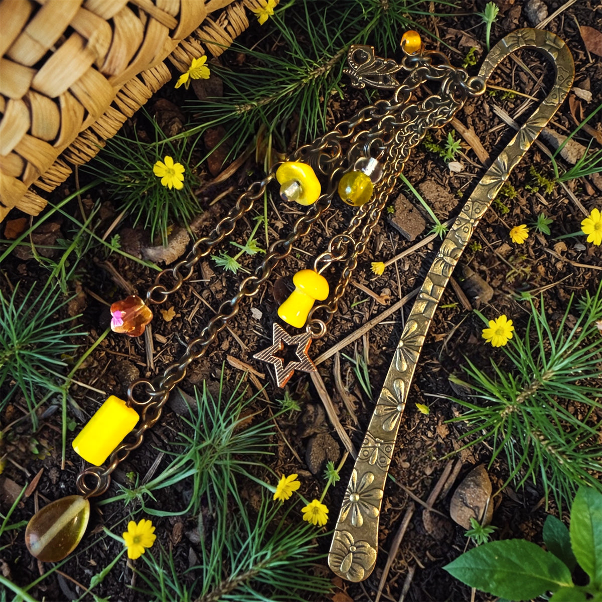 Antique brass charm bookmark with yellow glass beads, star charm, and woodland accents resting on forest floor with pine needles and small yellow flowers.